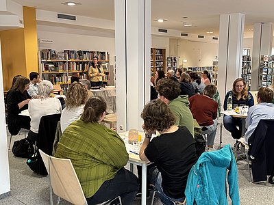 Publikum an mehreren kleinen Tischen lauscht einer Polizistin
