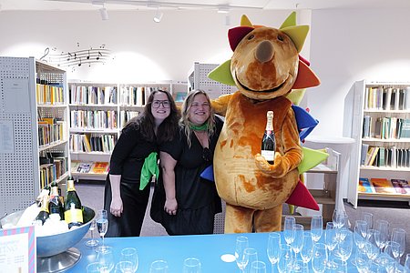 Zwei Frauen stehen neben einem Maskottchen mit Champagnerflasche in der Hand